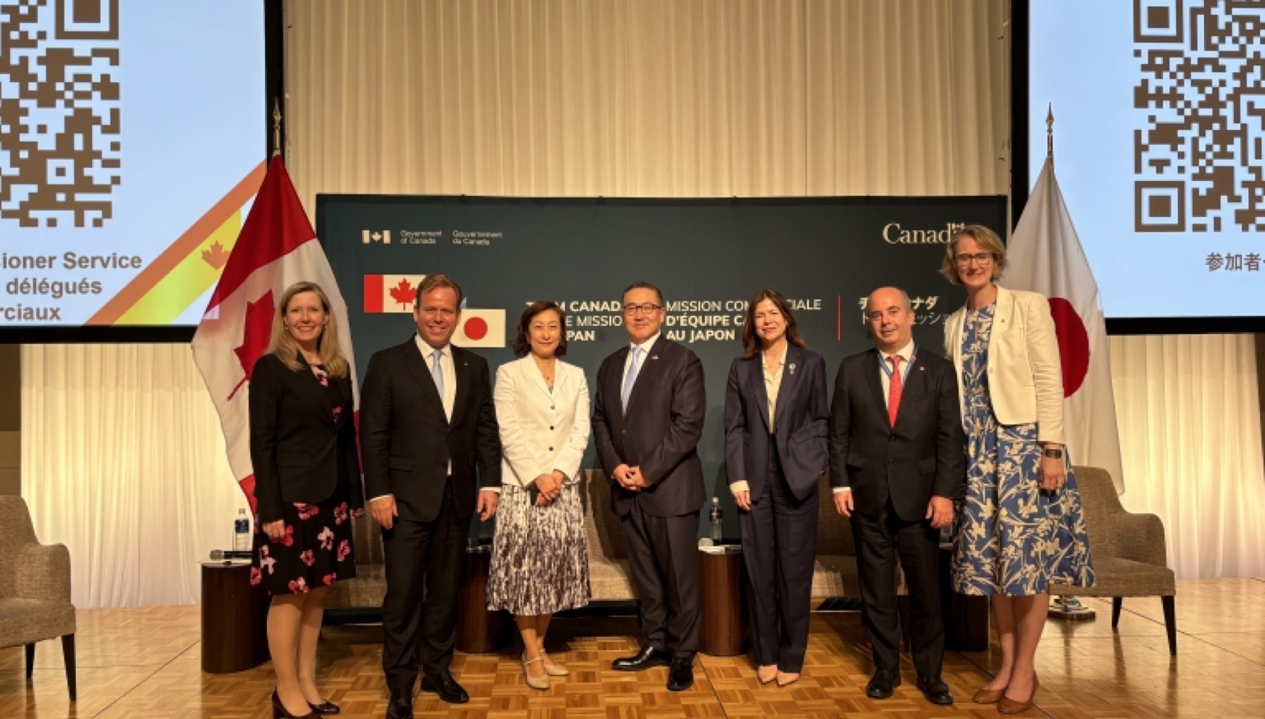 A delegation on stage between the Canadian and Japanese flags at the Team Canada Trade Mission